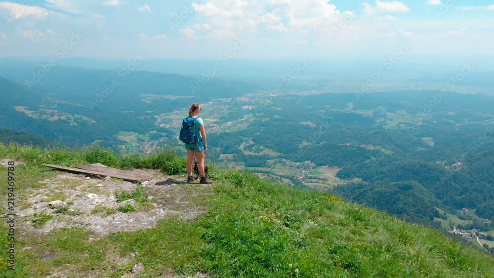 AERIAL: Female tourist looking at green valley after hiking up a grassy mountain