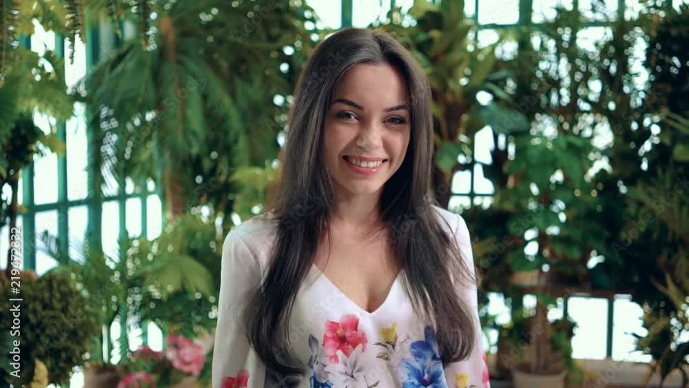 Smiling young girl with long dark hair looking at camera standing in the woods or a park on a sunny summer day. Left to right pan slow motion medium shot