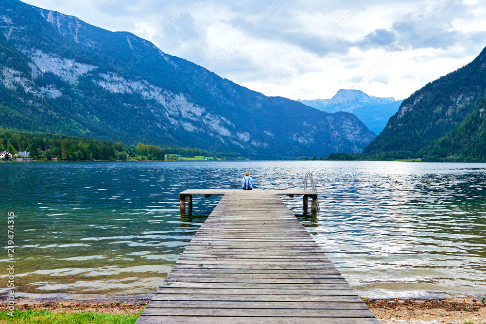 Self reflection. Woman sits on a wooden pier. Cloudy above the lake Hallstatt. 