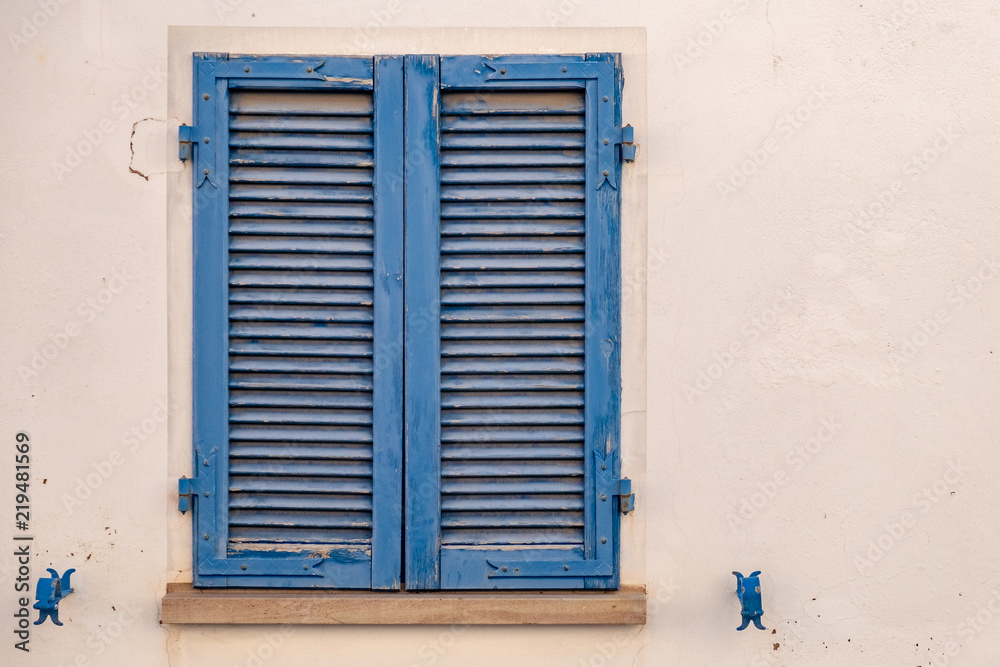 The window is covered with blue wooden shutters on the facade of the beige house