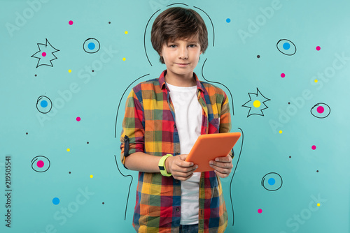 Modern tablet. Calm teenage boy looking glad and smiling while standing with his modern tablet after getting it for his birthday