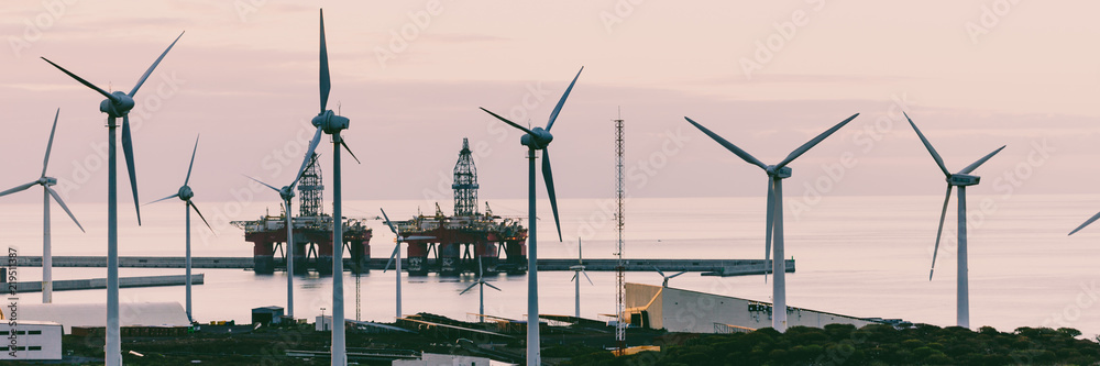 Group of windmills and solar panels for electric power production and oil rigs on coast.