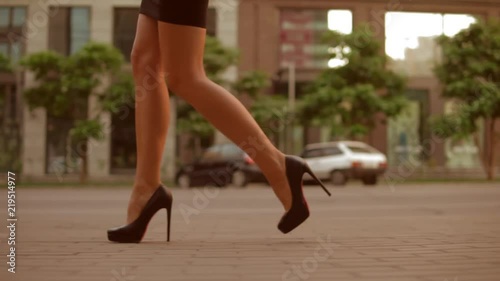 close up details businesswoman wearing black skirt and high-heeled shoes walking along the sidewalk. on the background buildings with storefront and cars parked along the road. slow motion