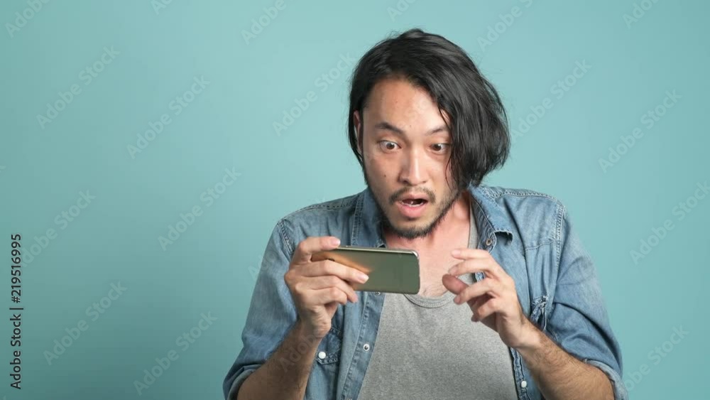 Young bearded male hipster looking at his phone to find great new from mobile message application. Bored Asian man looking at mobile phone and extremely happy as he see good news with blue background. Young bearded male hipster looking at his phone to find great new from mobile message application. Bored Asian man looking at mobile phone and extremely happy as he see good news with blue background.