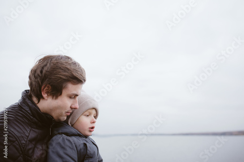 Father and son sitting at the lake bank in cold autumn