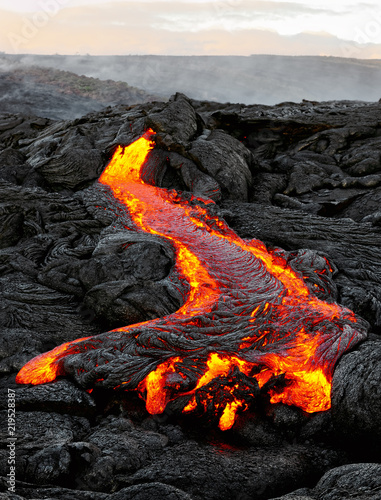 A lava flow emerges from an earth column and flows in a black volcanic landscape, in the sky shows the first daylight - Location: Hawaii, Big Island, volcano 