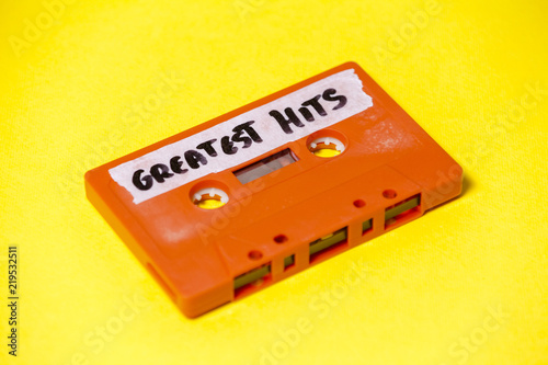 A vintage cassette tape (obsolete music technology), orange on a yellow surface, angled shot, carrying a label with the handwritten text Greatest Hits.
 photo