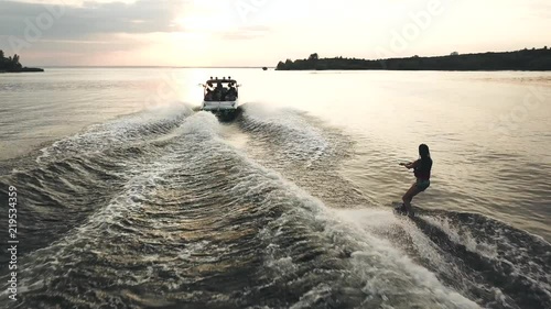 Flying over wakeboarder surfing behind boat. Aerial view of wakeboarding on river at sunset.