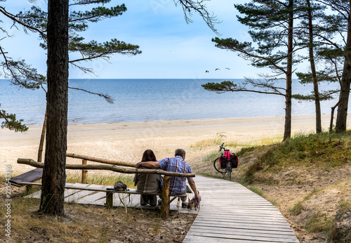 Happy couple of seniors are resting on wooden bench and looking at the distance on sandy beach of the Baltic Sea