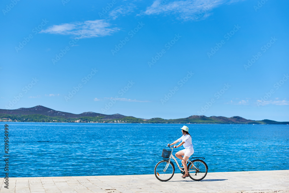 Beautiful female cyclist in white summer closing and hat riding bicycle along stony sidewalk on blue sea water and resort town at foot of mountains on opposite shore background. Tourism and vacations.