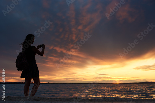 Silhouette of young tourist woman in short dress and with backpack and camera standing alone at water edge taking picture of beautiful seascape at sunset. Tourism and vacations concept.