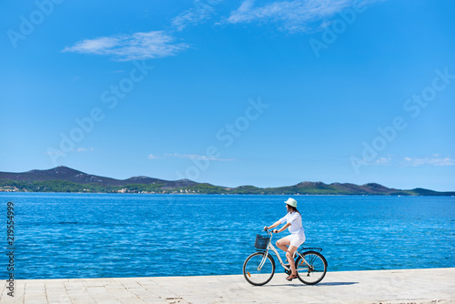 Beautiful female cyclist in white summer closing and hat riding bicycle along stony sidewalk on blue sea water and resort town at foot of mountains on opposite shore background. Tourism and vacations.