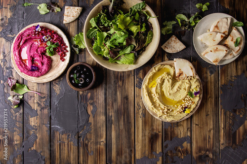 Variety of homemade traditional and beetroot spread hummus with pine nuts, olive oil, pomegranate served on ceramic plates with pita bread and green salad on dark wooden background. Flat lay, space.