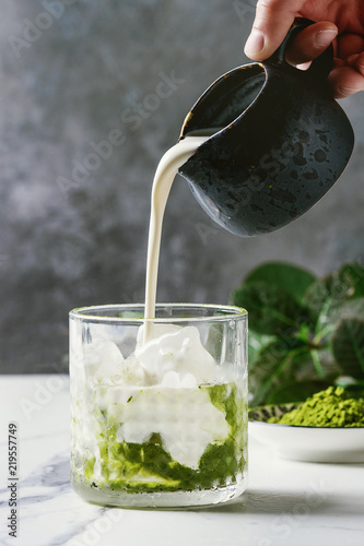 Cream pouring from jug to matcha green tea iced latte or cocktail in glass, with ice cubes, matcha powder on white marble table, decorated by green branches. Grey wall at background