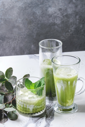 Matcha green tea iced latte or cocktail in three different glasses with ice cubes, matcha powder and jug of milk on white marble table, decorated by green branches. Grey wall at background