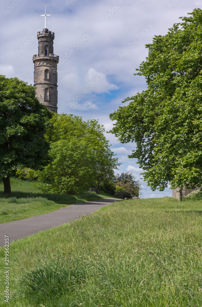Edinburgh, Scotland, UK - June 13, 2012: Nelson Monument on Calton Hill captured between green trees behind green lawn and under blue cloudy sky.
