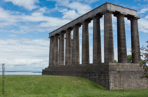 Edinburgh, Scotland, UK - June 13, 2012: Free standing National Monument is row of pillars under blue sky with white clouds. North Sea inlet in back, green grass in front.