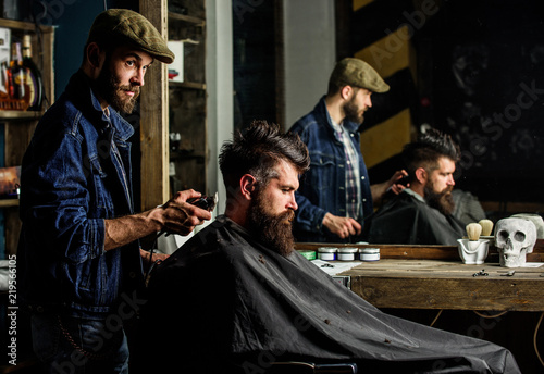 Barber styling hair of brutal bearded client with clipper. Barber with hair clipper works on hairstyle for bearded man barbershop background. Hipster client getting haircut. Hipster lifestyle concept