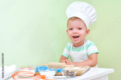 Cheerful cute baby boy in a chef costume smiling standing at the table where doing cookies