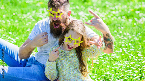 Rock star concept. Dad and daughter sits on grass at grassplot, green background. Child and father posing with star shaped eyeglases photo booth attribute at meadow. Family spend leisure outdoors. photo