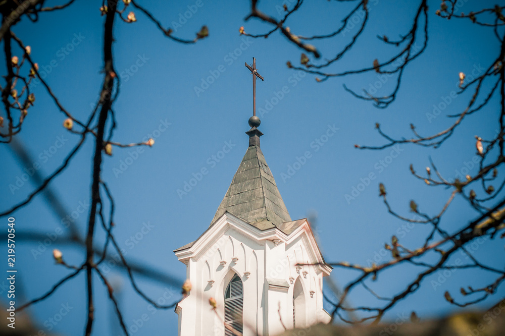 View of the tower of a Christian church with a cross through the branches of a tree View of the tower of a Christian church with a cross through the branches of a tree