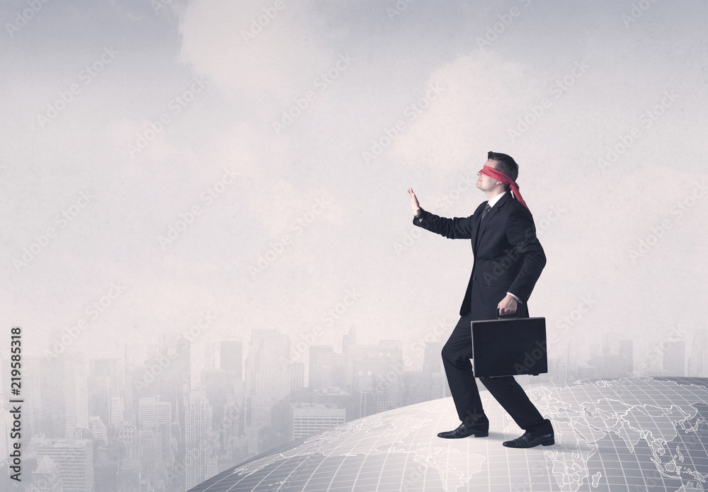 Young blindfolded businessman steps on a grey world map with a city in the background Young blindfolded businessman steps on a grey world map with a city in the background