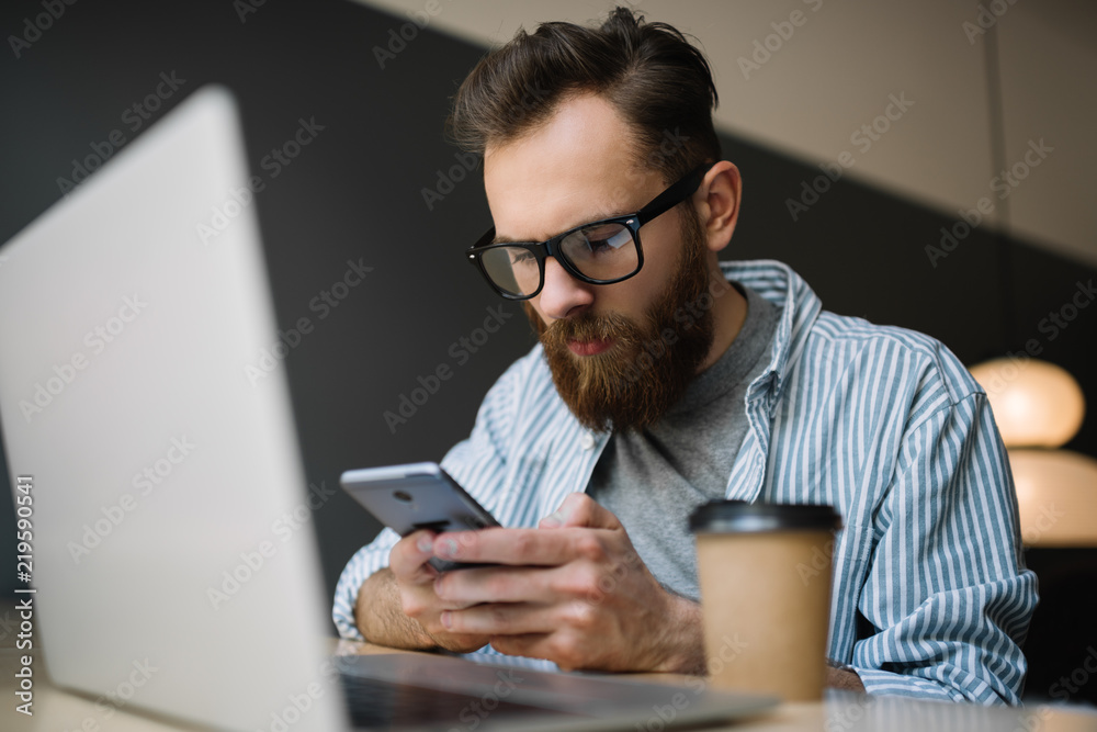 Bearded man wearing hipster glasses using smartphone and laptop computer for his project, downloading application, researching information online. Young caucasian freelancer working in modern office. Bearded man wearing hipster glasses using smartphone and laptop computer for his project, downloading application, researching information online. Young caucasian freelancer working in modern office.