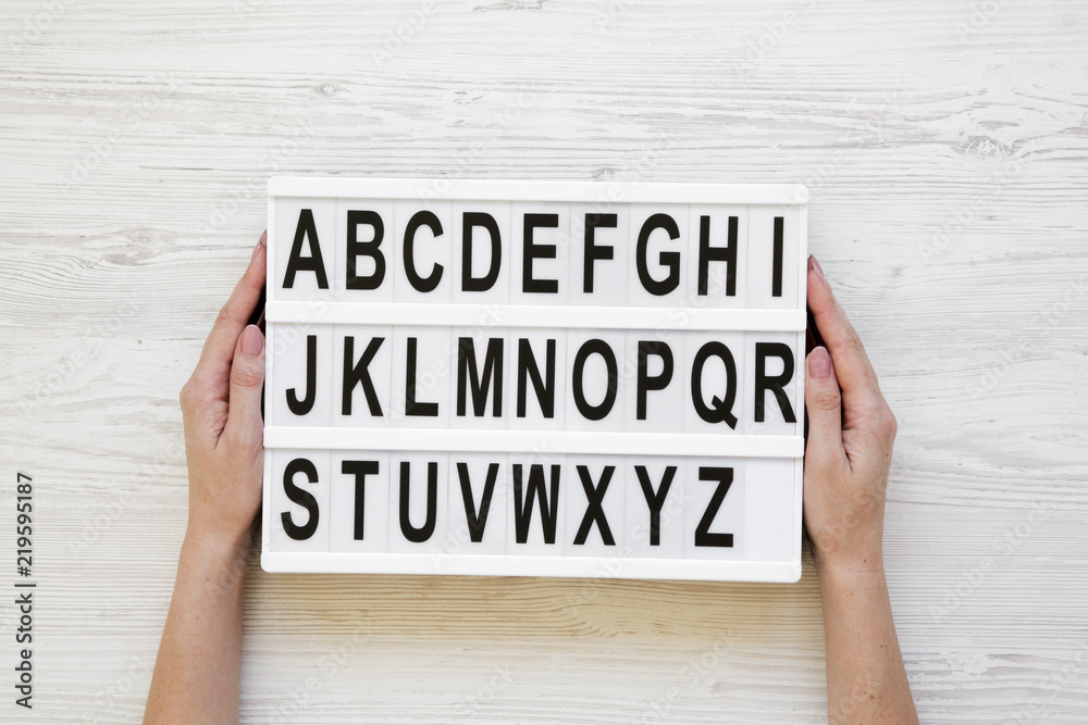 Female hands hold modern board with english alphabet on lightbox over white wooden background, from above. Closeup. Female hands hold modern board with english alphabet on lightbox over white wooden background, from above. Closeup.