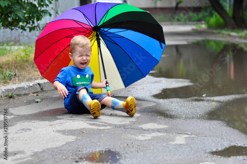 A two year old cute boy cries and sits under a multicolored rainbow umbrella photo