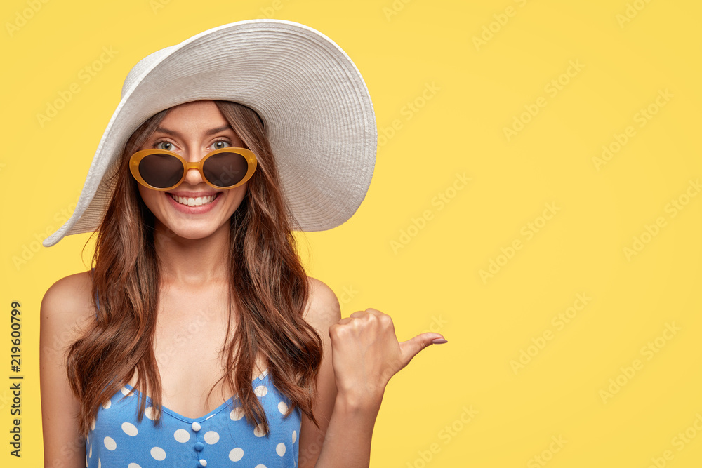 Summer time concept. Cheerful beautiful brunette woman in stylish sunglasses and hat, has friendly smile, points with thumb aside, shows blank space for your advertisement or promotional text - obrazy, fototapety, plakaty 