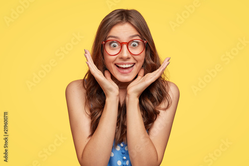 Photo of satisfied overjoyed woman smiles broadly, cant believe in her success, stares through spectacles, stands alone in studio against yellow background. People, facial expressions and feeling