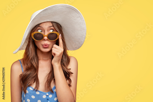 Stylish woman with surprised mysterious expression, looks asdie, wears stylish summer shades, hat and polka dot blouse, isolated over yellow background with copy space for your text. Relaxation