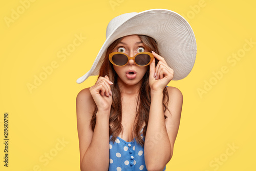 Fashionable lady wears summer hat and sunglasses, expresses panic and fear, stares with shock at camera, surprised have spoiled vacation, isolated over yellow background. Seasonal holidays concept