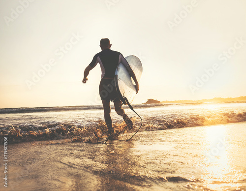Surfer with surfboard runs in ocean waves, sunset time. Active lifestyle concept