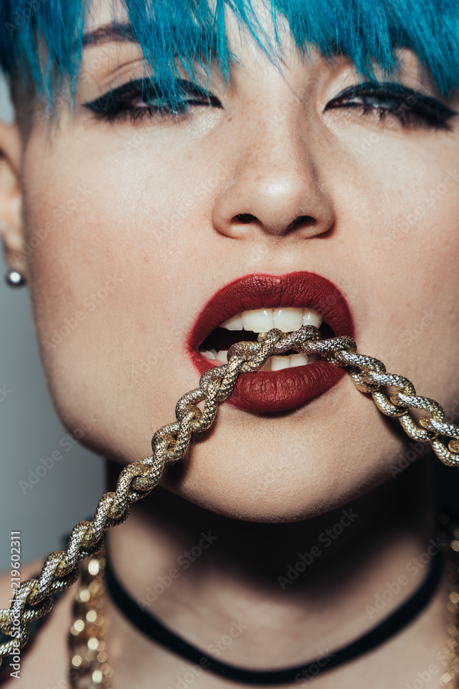 Close up fashion portrait photography of a girl with blue hair on a white background She bites a thick yellow chain. Professional model posing in studio.