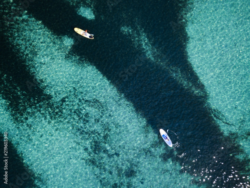 Aerial view of two people doing stand up paddleboarding (SUP) on a beautiful turquoise sea. Cala Brandinchi, Sardinia, Italy.