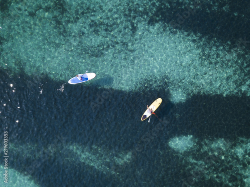 Aerial view of two people doing stand up paddleboarding (SUP) on a beautiful turquoise sea. Cala Brandinchi, Sardinia, Italy.