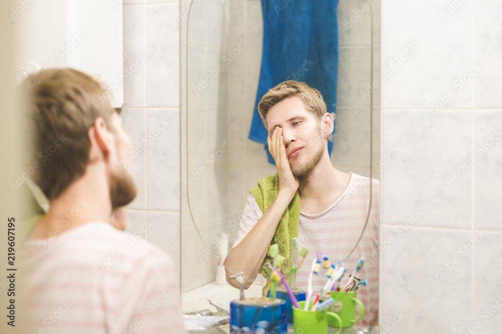 Fototapeta premium young bearded man with towel looking into mirror in bathroom, sleepy morning