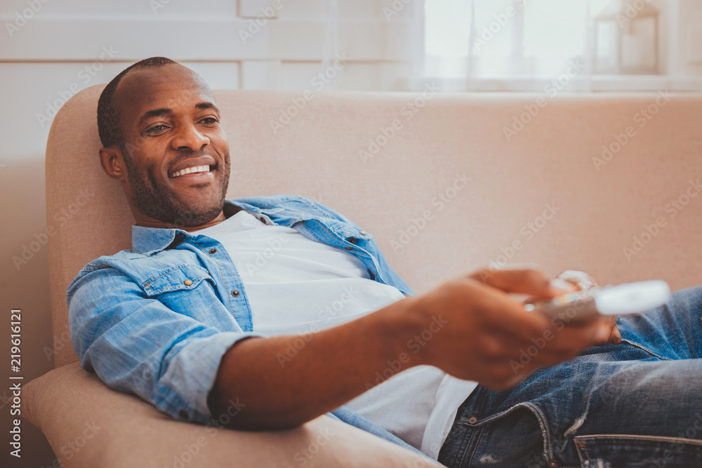 Fototapeta premium My weekend. Joyful afro-american man lying on the sofa and holding a remote control