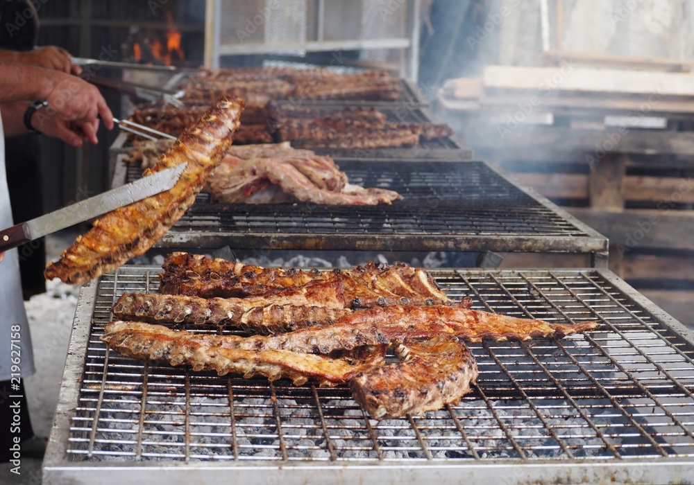 Large barbecue of grilled pork ribs with tongs and knives, on the background, that turn the meat on the grill,