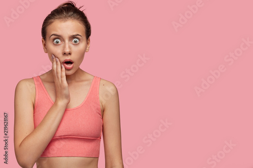 Stupefied young sportswoman with healthy skin, stares with shock, finds out about loosing sport competition, dressed in top, isolated over pink background with copy space for your advertisment photo