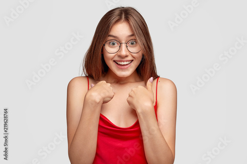 Me, really? Cheerful young cute woman with unexpected expression points at herself, being surprised to recieve words of praise, dressed casually, has joyful look, poses against white background.