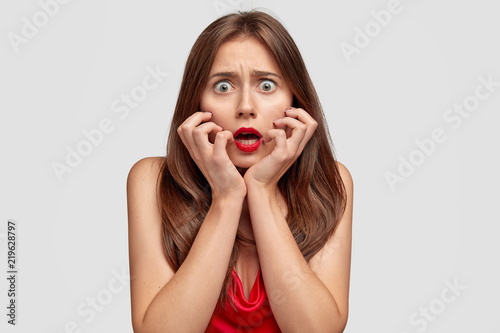 Fearful nervous woman keeps hands on cheeks, stares with bugged eyes, opens mouth from fright, has red lips, poses against white background. Shocked stupefied wife notices husband with lover