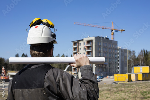Construction worker in dirty overalls and a white helmet in Finland. In his hand he has drawings. In the background is out of focus in the construction site. 