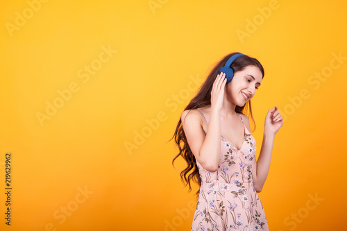 Cute young woman listening music on her headphones in studio over yellow background.