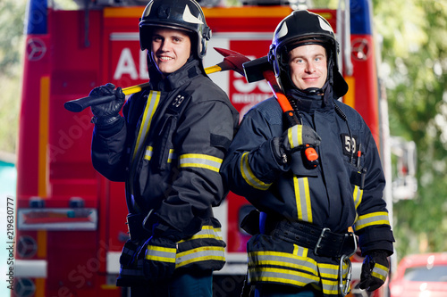 Photo of two young firemen with axes in hands near fire engine © Sergey