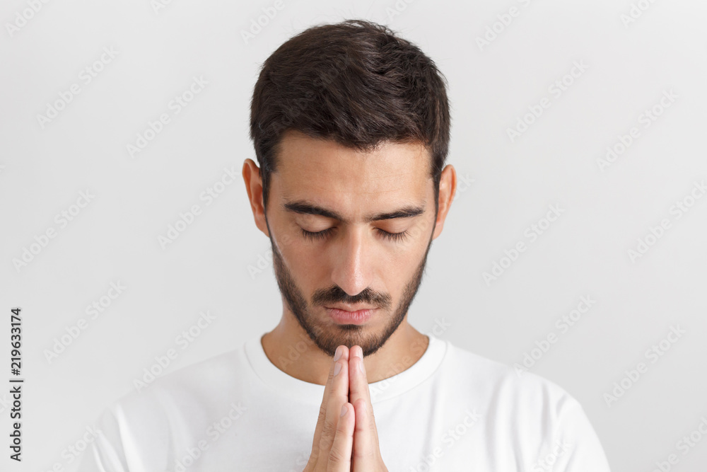 Daylight shot of young man in white tshirt isolated on gray background looking stressed, putting hands together as if he is praying with closed eyes