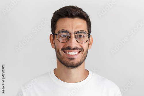 Daylight portrait of young handsome caucasian man isolated on grey background, dressed in white t-shirt and round eyeglasses, looking at camera and smiling positively photo