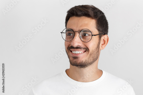 Indoor portrait of handsome young european caucasian man isolated on gray background  dressed in casual white t-shirt  wearing round glasses  having turned aside as if looking at something