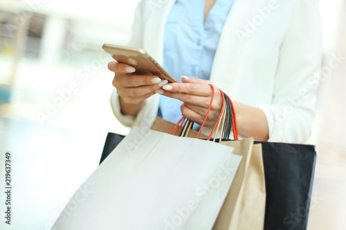 Woman using smartphone and holding shopping bag while standing on the mall background.